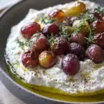 Whipped feta dip with roasted grapes - the image is a close-up of a dish in a gray bowl. the dish appears to be a type of hummus or dip, with a creamy white filling and a layer of red and yellow olives on top. the olives are arranged in a circular pattern, with some of them overlapping each other. there are also some sprigs of thyme scattered on top, adding a pop of green color to the dish. the bowl is sitting on a wooden table, and there is a wooden spoon resting on the side of the bowl.