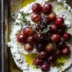Whipped feta dip with roasted grapes - the image shows a rectangular baking dish filled with a creamy white substance, which appears to be ricotta or feta cheese. on top of the ricotta, there are several red cherries arranged in a circular pattern. the cherries are garnished with a sprig of thyme, adding a pop of green color to the dish. the dish is sitting on a dark wooden surface, and the overall color scheme of the image is warm and inviting.