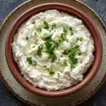 Whipped feta goat cheese dip - the image is a close-up of a bowl of mashed potatoes. the bowl is made of terracotta and has a brown rim with a speckled pattern. the mashed potatoes are creamy and smooth, and are topped with a generous amount of chopped parsley. the background is a dark grey textured surface, and there are a few sprigs of parsley scattered around the bowl. the overall mood of the image is rustic and appetizing.