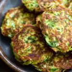 Zucchini feta fritters - the image shows a plate of zucchini fritters on a wooden table. the plate is made of dark brown ceramic and has a textured surface. the zucchinis are golden brown and appear to be freshly cooked. they are arranged in a circular pattern on the plate, with some overlapping each other. there are also some dill leaves scattered around the plate. the background is blurred, but it appears to be a rustic kitchen countertop.