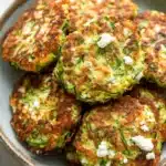 Zucchini feta fritters - the image shows a plate of zucchini fritters. the plate is made of ceramic and has a light brown color. the fritter in the center of the plate is golden brown and appears to be crispy on the outside. it is garnished with chopped herbs and crumbled feta cheese. the zucchinis are arranged in a circular pattern on the plate, with some overlapping each other. the background is white, making the plate and its contents stand out.